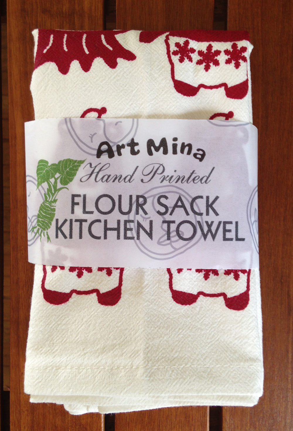  A white flour sack kitchen towel with red hand-painted designs. The towel is folded and has a label that reads 'Art Mina Hand Printed Flour Sack Kitchen Towel'.